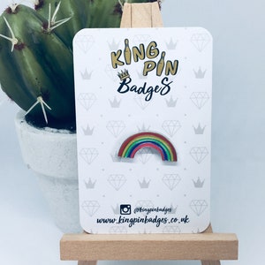 May include: Rainbow enamel pin on a white card with the text "King Pin Badges" and a crown. The card has a repeating pattern of crowns and diamonds. The text "@kingpinbadges" and "www.kingpinbadges.co.uk" is also on the card.
