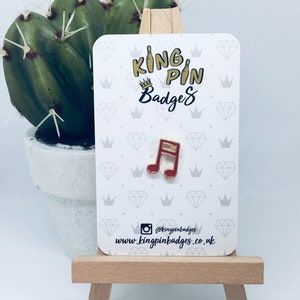 Op de afbeelding: Een rode en gouden emaille muzieknootspeld op een witte kaart met de tekst "KING PIN Badges". De kaart is versierd met diamant- en kroongrafieken. De speld wordt gepresenteerd op een klein houten ezel.