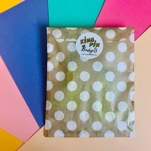 May include: A brown paper bag with white polka dots. The bag has a sticker on it that says "King Pin Badges" with a crown above the word "King".