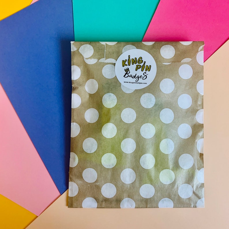 May include: A brown paper bag with white polka dots. The bag has a sticker on it that says "King Pin Badges" with a crown above the word "King".