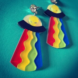 May include: A pair of rainbow-colored alien spaceship earrings. The earrings are made of acrylic and feature a black spaceship with a yellow alien inside. The spaceship is surrounded by a rainbow-colored halo.