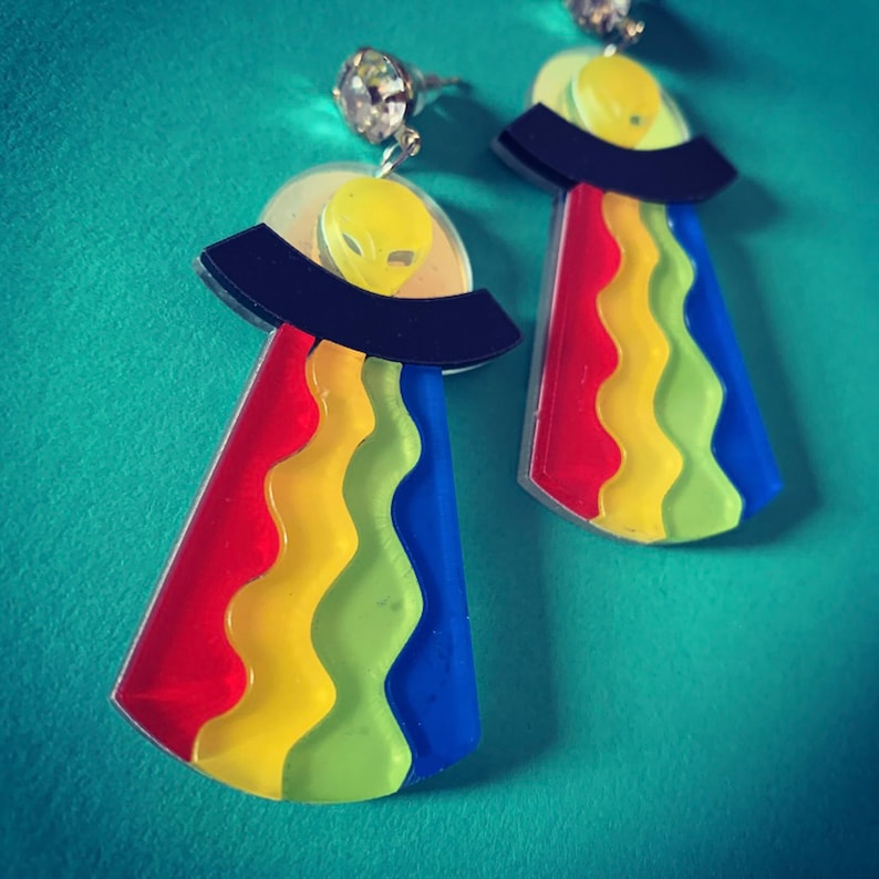 May include: A pair of rainbow-colored alien spaceship earrings. The earrings are made of acrylic and feature a black spaceship with a yellow alien inside. The spaceship is surrounded by a rainbow-colored halo.