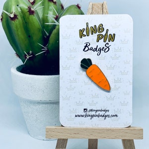 May include: A carrot shaped enamel pin on a white card with the text "King Pin Badges" and the website address "www.kingpinbadges.com".