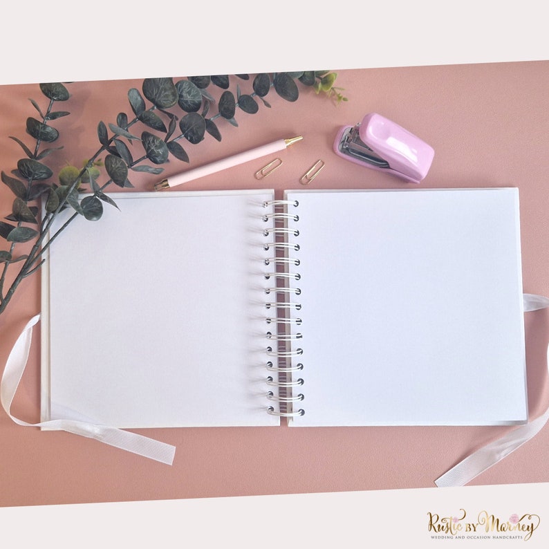 Peut inclure: Un carnet blanc &agrave; spirales ouvert avec un ruban blanc en guise de marque-page. Le carnet est sur une surface rose avec une agrafeuse rose et un stylo.