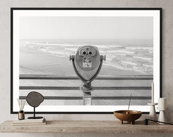 May include: Black and white photograph of a vintage coin-operated telescope on a pier overlooking a beach with waves crashing in the distance.