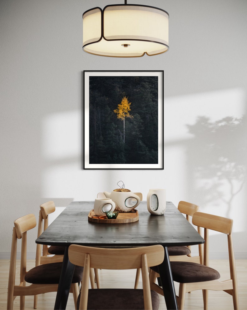 May include: A black framed print of a single yellow tree in a dark forest setting hangs above a dark wood dining table with six chairs. The table is set with a teapot and two mugs.