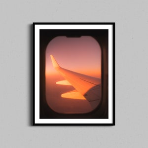 Pode incluir: Uma vista da janela de um avião, mostrando a asa do avião contra um céu de pôr do sol laranja e rosa vibrante.