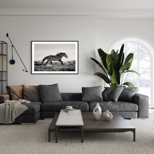 May include: A black and white photograph of a horse running through a field, framed and hanging on a white wall in a living room. The room features a gray sectional sofa, a coffee table, a potted plant, and a window with a view of a snowy landscape.