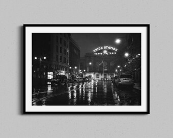 May include: Black and white photograph of a framed print. The image depicts a city street at night, with the words "UNION STATION TRAVEL TRAIN" illuminated above a building. The street is wet, reflecting the lights.