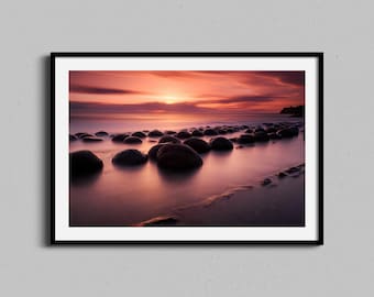 May include: A photograph of a sunset over the ocean with large, round rocks in the foreground. The sky is a vibrant orange and pink, and the water is a deep blue. The rocks are smooth and grey, and they are partially submerged in the water.