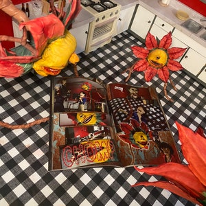 May include: Three red and yellow flower-like creatures with toothy grins are crawling on a black and white checkered floor. A comic book with a similar creature on the cover is open on the floor. The comic book has the word "CHOMP" on the cover.