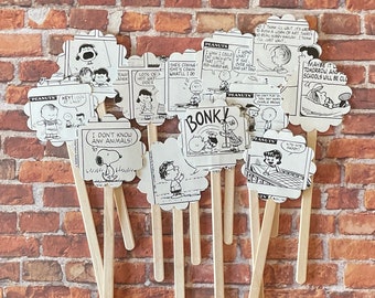 May include: A collection of Peanuts comic strip cutouts on wooden sticks. The black and white images feature various characters and speech bubbles with phrases like "BONK!" and "I don't know any animals!" against a brick background.