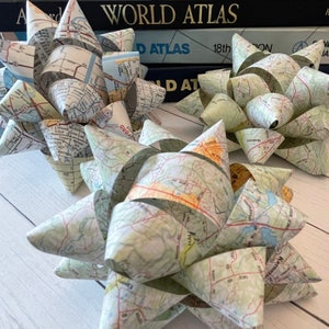 May include: Two handmade paper bows crafted from vintage map pages. The bows are star-shaped, with multiple layers of folded paper creating a textured, dimensional effect. The maps feature a mix of green, beige, and orange hues, with visible road and city details.