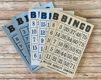 May include: A collection of vintage bingo cards. The cards are in shades of blue and off-white, with black text and numbers. The cards are fanned out on a wooden surface. The text "BINGO" is prominently displayed.