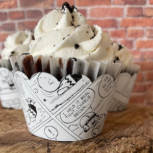 May include: Three cupcakes with white frosting and chocolate sprinkles sit on a wooden surface. The cupcake liners feature a black and white comic strip design with text that reads "I held it real tight, Charlie Brown..." and "I'm going for the day."