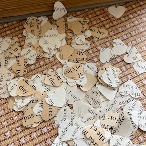 May include: A collection of heart-shaped paper confetti scattered on a patterned surface. The confetti is made from book pages, with visible text. Some hearts are off-white, others are tan.