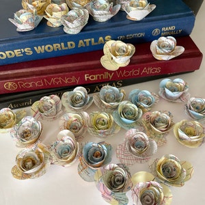 May include: A collection of paper flowers made from vintage maps. The flowers are in various shades of blue, yellow, and cream. The flowers are arranged on a white surface.