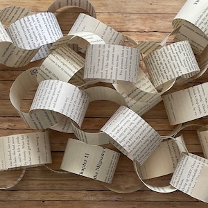 May include: A paper chain made from pages of a book. The chain is made of individual loops of paper, each with text from the book printed on it. The chain is lying on a wooden surface.
