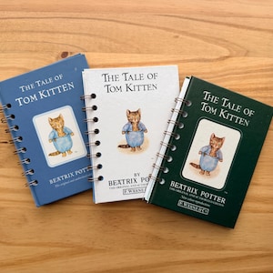May include: Three spiral-bound books titled "The Tale of Tom Kitten" by Beatrix Potter. The books are in blue, white, and green, each featuring an illustration of a cat in a blue outfit. The books are on a wooden surface.