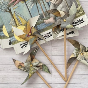 May include: Three decorative pinwheels with patterned paper blades and wooden sticks are arranged in front of several books. The pinwheels feature various colors and designs, including floral and geometric patterns. The books have the title "STORIES PICTURES MAURICE SENDAK" printed on the cover.