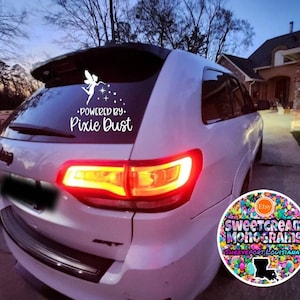 May include: White car window decal with a fairy graphic and the words "Powered by Pixie Dust." The decal is on the rear window of a white SUV. The image also includes a colorful Etsy shop logo.