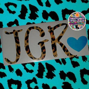 May include: A white decal featuring the initials "JGK" in a leopard print design, with a light blue heart. The decal is set against a teal background with black leopard spots. A circular logo is in the upper right corner.