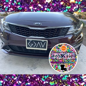 May include: A dark brown Kia car with a license plate that reads "G>AV". The car is parked in front of a colorful background with a round sticker that says "Sweetcream Monograms Shreveport, Louisiana" and has a small Etsy logo on it.