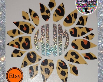May include: A vinyl decal featuring a sunflower with a leopard print design. The center of the sunflower has a silver glitter monogram with the letters "KLM".