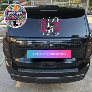 May include: A vinyl decal applied to the rear window of a black SUV. The decal features a monogram with the letters B and N in a bright pink script font, flanking a central letter H with a brown and white cow print pattern.