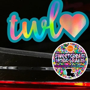 May include: A turquoise blue vinyl decal with the word "two" in a rainbow iridescent font and a heart with a rainbow iridescent finish. The decal is on a black car. The decal is from Sweetcream Monograms, Shreveport, Louisiana.