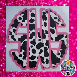 May include: A pink and white cow print monogram with a scalloped edge. The monogram is in the shape of a circle and features the letters "S W E".