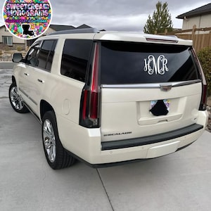 May include: White Cadillac Escalade SUV with a monogrammed "H.N.C.R." decal on the rear window. The vehicle is parked on a concrete driveway.