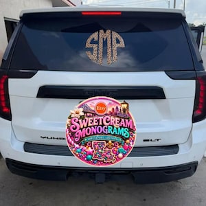 May include: A white SUV with a circular leopard print monogram decal featuring the letters SMP applied to the rear window. The vehicle has black trim, vertical red taillights, and a GMC emblem on the rear hatch above a colorful circular logo.