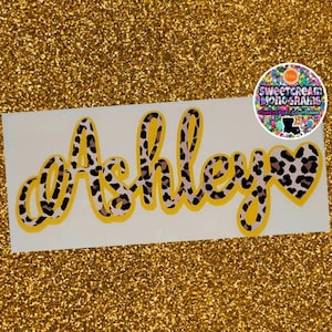 May include: A yellow and brown leopard print vinyl decal with the name "Ashley" and a heart.
