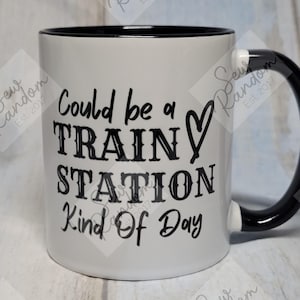 May include: White ceramic mug with a black interior and handle. The mug has the text "Could be a TRAIN STATION Kind Of Day" in black, with a heart symbol. The handle has a black and white striped design.