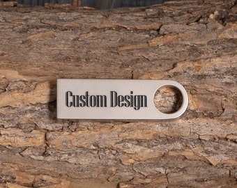 Puede incluir: Una memoria USB de metal plateado con el texto "Custom Design" grabado en ella. La memoria está sobre un trozo de corteza de árbol marrón.