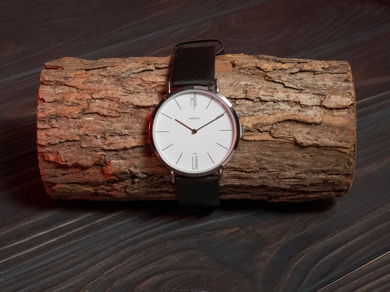 May include: A silver watch with a white face and black leather band. The watch is on a piece of wood.