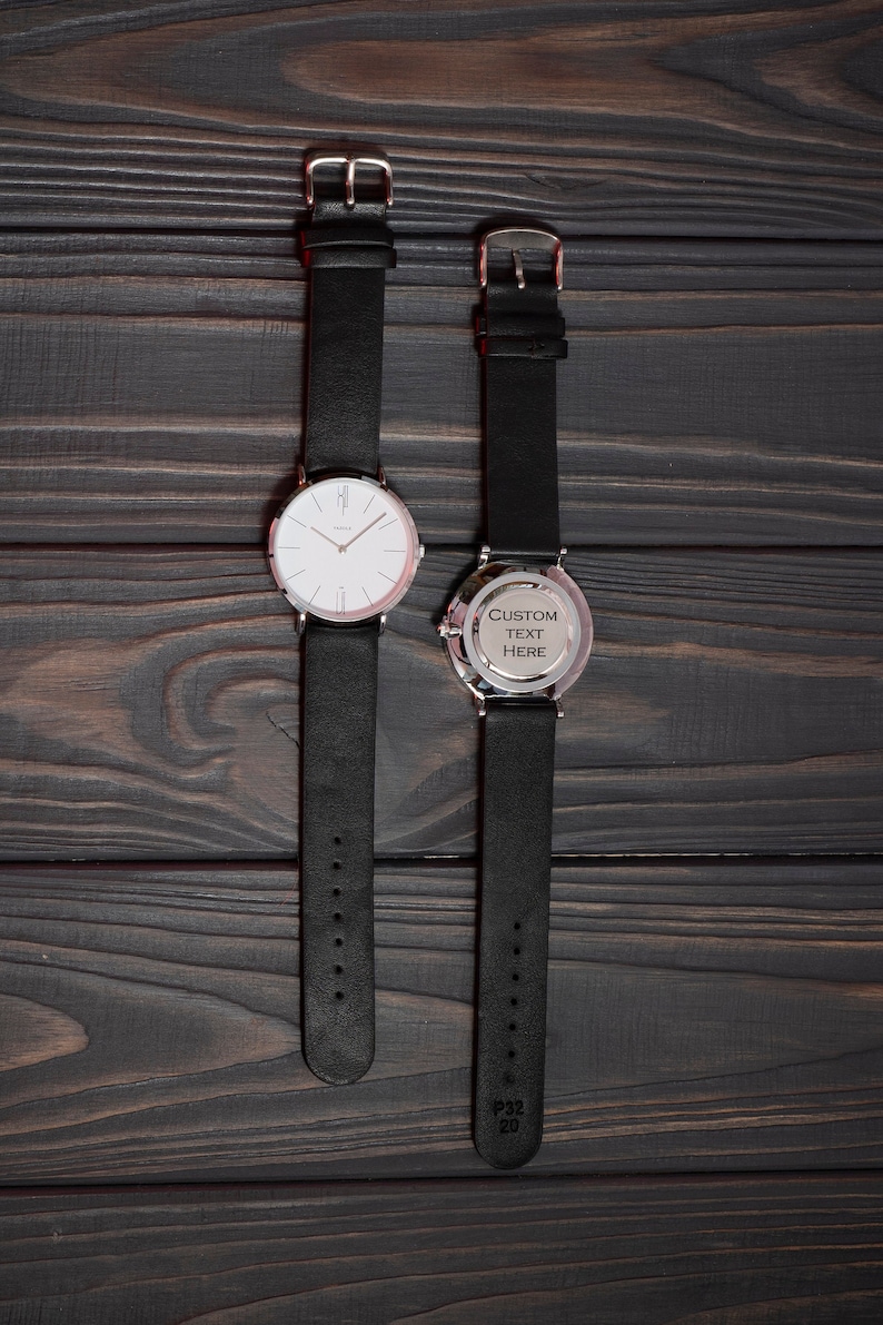 May include: Two silver wristwatches with black leather bands on a dark wood background. The watch on the left has a white face with silver hands and markers. The watch on the right has a silver face with the text "CUSTOM TEXT HERE" engraved on it.