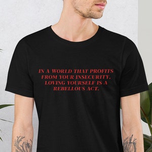 Könnte beinhalten: Ein schwarzes T-Shirt mit rotem Text, der "IN A WORLD THAT PROFITS FROM YOUR INSECURITY, LOVING YOURSELF IS A REBELLOUS ACT." lautet.