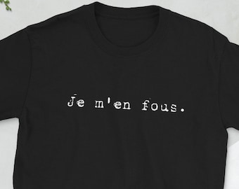 Op de afbeelding: Zwart T-shirt met korte mouwen met de witte tekst "Je m'en fous". Het shirt wordt gepresenteerd op een licht oppervlak, met een camera, portemonnee, telefoon en bril in de buurt. Een takje groen is ook zichtbaar.