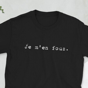 Puede incluir: Camiseta negra de manga corta con la frase "Je m'en fous" impresa en blanco. La camiseta se muestra sobre una superficie clara, con una cámara, una cartera, un teléfono y gafas cerca. También es visible una ramita de vegetación.