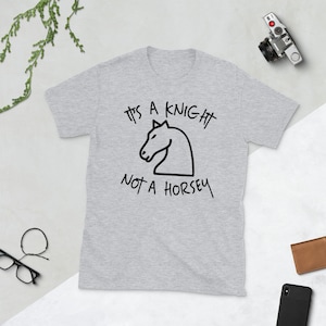 Puede incluir: Una camiseta gris con un gráfico negro de un caballero de ajedrez y el texto "It's a knight not a horsey".