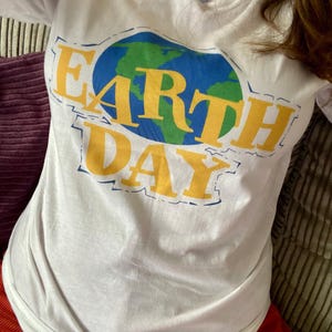 May include: White long-sleeve t-shirt with a graphic that says "EARTH DAY". The graphic features a blue and green globe with yellow lettering. The shirt is made of a soft material.