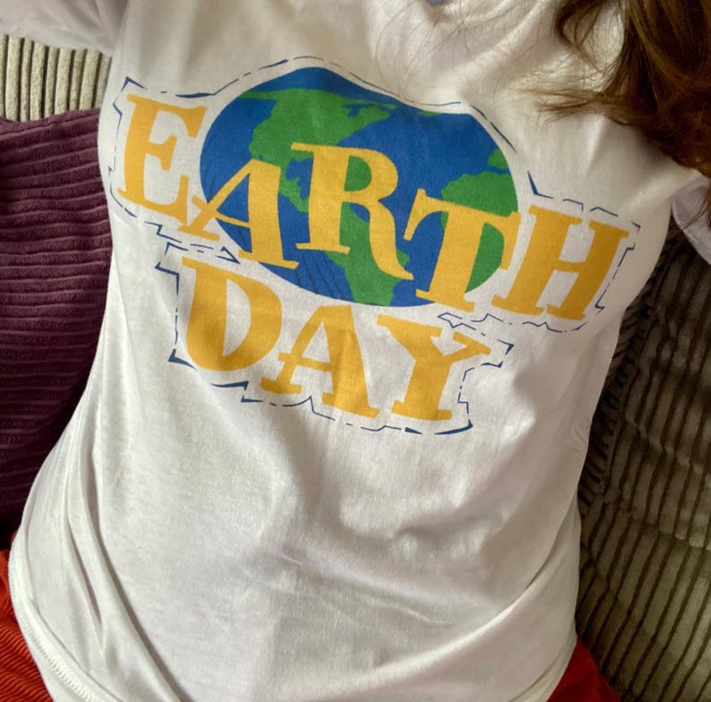 May include: White long-sleeve t-shirt with a graphic that says "EARTH DAY". The graphic features a blue and green globe with yellow lettering. The shirt is made of a soft material.