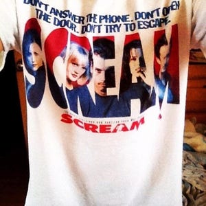May include: White t-shirt featuring the word "SCREAM" in red and blue, with a graphic of characters from the movie. The shirt also has the text "DON'T ANSWER THE PHONE. DON'T OPEN THE DOOR. DON'T TRY TO ESCAPE."