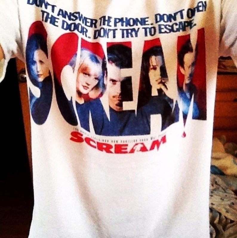 May include: White t-shirt featuring the word "SCREAM" in red and blue, with a graphic of characters from the movie. The shirt also has the text "DON'T ANSWER THE PHONE. DON'T OPEN THE DOOR. DON'T TRY TO ESCAPE."