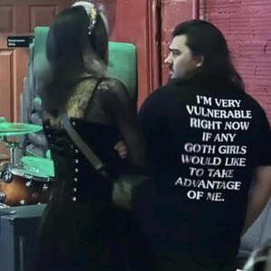 May include: A black t-shirt with white text that reads "I'M VERY VULNERABLE RIGHT NOW IF ANY GOTH GIRLS WOULD LIKE TO TAKE ADVANTAGE OF ME." The shirt is worn by a person with long dark hair. Another person in a black dress is also in the image.