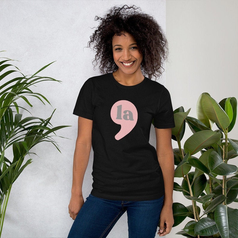 Puede incluir: Una camiseta negra con un punto y coma rosa y la palabra "la" en gris.