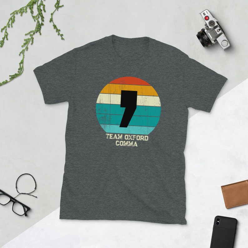 May include: A dark grey t-shirt with a retro design. It features a black comma inside a circle with orange, yellow, teal, and cream stripes. The text "TEAM OXFORD COMMA" is below. A camera, glasses, and wallet are in the background.