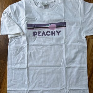 May include: White t-shirt with a graphic design featuring a peach illustration and the word "PEACHY" in purple letters. The design includes a horizontal stripe in shades of purple and brown.